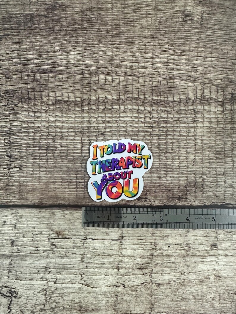 May include: A colorful sticker with the text "I told my therapist about you." The text is in a rainbow tie-dye design. A metal ruler is next to the sticker, showing inches.