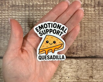 May include: A white sticker with the text "EMOTIONAL SUPPORT" above a cartoon quesadilla slice. The quesadilla is yellow with a smiling face and dripping cheese. The text "QUESADILLA" is below the image.