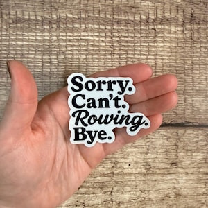 May include: A white sticker with black text that reads "Sorry. Can't. Rowing. Bye." The sticker is in a cursive font and is on a wooden surface.