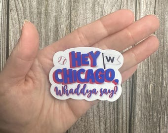 May include: White sticker with a red and blue baseball themed design. The sticker says "Hey Chicago, Whaddaya say?" with a baseball graphic and a "W" for win.