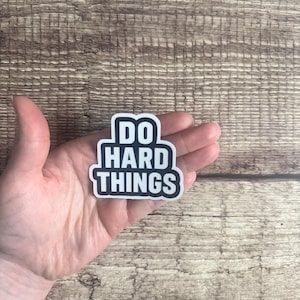 May include: A black and white sticker with the text "DO HARD THINGS" in a bold, sans-serif font. The sticker has a white border and is held in a hand against a wooden background.