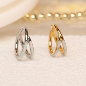 Op de afbeelding: Twee oorringen, één zilver en één goud, elk met een witte opaal inleg. De oorbellen hebben een dubbel-hoepel ontwerp. De achtergrond is een zacht wit oppervlak met een vage gouden ketting.