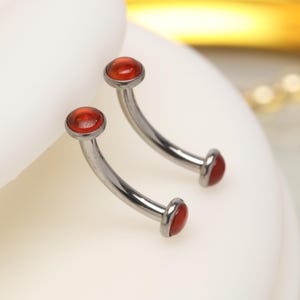 Puede incluir: Un par de piercings curvos para cejas. Cada anillo tiene una barra curva de color plateado con una pequeña gema roja y redonda en cada extremo. Las joyas se exhiben sobre una superficie blanca.