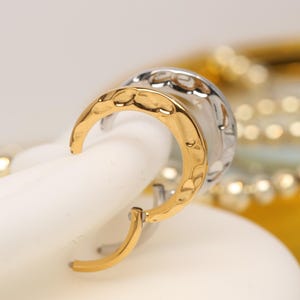 May include: A pair of gold and silver hoop earrings. The gold earring has a hammered texture, while the silver earring has a textured design. The earrings are displayed on a white surface, with a blurred background.