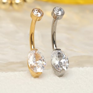 May include: Two belly button rings. One is gold-toned, the other silver-toned. Each has a curved bar with a small, round, clear gem at the top and a larger, oval, clear gem at the bottom. The gems sparkle, reflecting light.