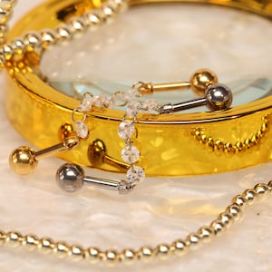 Puede incluir: Primer plano de joyería corporal, que incluye pendientes de barra con bolas doradas y plateadas. Una lupa dorada y un collar de cuentas también están en la imagen. Las joyas se exhiben sobre una superficie blanca.