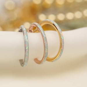 May include: Three hoop earrings in silver, rose gold, and gold, each with a white opal inlay. The earrings are displayed on a white cylindrical surface, with a soft, blurred background.