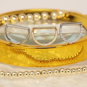 Könnte beinhalten: Drei Septumringe auf einer goldenen Oberfläche. Einer ist silberfarben, einer goldfarben mit einem opalartigen Einsatz und der dritte silberfarben mit kleinen, klaren Steinen. Die Ringe haben ein Halbkreis-Design mit einem geraden Balken oben.