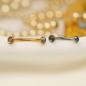 May include: Two barbell-style body jewelry pieces. One is gold-toned with light green gemstones, the other is silver-toned with dark green gemstones. Both have a straight bar design with a gemstone at each end.