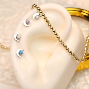 Könnte beinhalten: Vier silberne Halbmond-Ohrringe mit verschiedenen Edelsteinen in Farbe. Die Ohrringe werden am Ohr eines Modells gezeigt. Die Ohrringe sind eine beliebte Wahl für Knorpelpiercings.