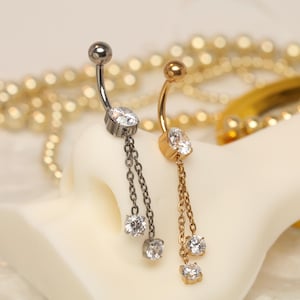 Puede incluir: Dos piercings para el ombligo. Uno es plateado con una cadena colgante de tres gemas redondas y transparentes. El otro es dorado con una cadena de gemas similar. Ambos tienen un diseño de barra curva y se muestran sobre una superficie blanca.