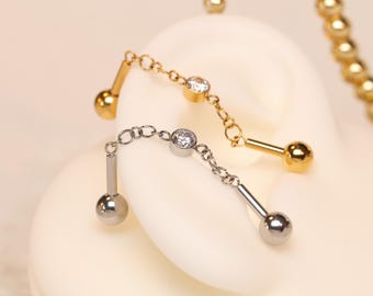 Op de afbeelding: Close-up van twee gouden en zilveren kettingoorbellen. Elke oorbel heeft een kleine, ronde, heldere edelsteen en een bungelende ketting met een bal aan het uiteinde. De oorbellen worden gepresenteerd op een wit oppervlak.