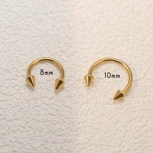 May include: Two gold-coloured, horseshoe-shaped, body piercing rings with pointed ends. The ring on the left is 8mm in diameter and the ring on the right is 10mm in diameter.
