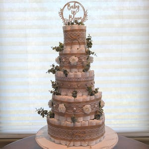 May include: A multi-tiered diaper cake, decorated with burlap, lace, and white roses. The cake is topped with a wooden sign that reads "Baby in Bloom". Greenery accents the cake, which sits on a white, beaded cake stand.