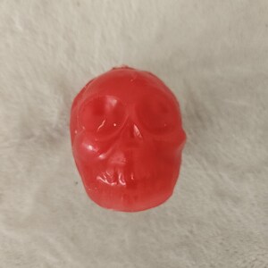 May include: A small, red skull-shaped candle. The candle has a smooth, glossy finish and is resting on a soft, white surface. The skull's features, including eye sockets, nose, and teeth, are clearly defined. This decorative candle is perfect for Halloween.