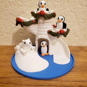 May include: A white ceramic penguin playset with a polar bear, a slide, and a tower decorated with green garland and red berries.