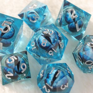 May include: A set of seven polyhedral dice in various shapes. The dice are transparent blue with a detailed eye design inside. Silver numbers are visible on each face, creating a fantasy gaming theme.