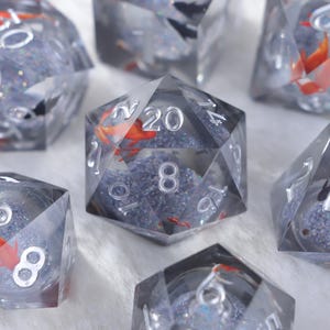 May include: Set of clear resin dice with silver numbering and a goldfish design. The dice are filled with a shimmering, glittery substance and small orange fish. The dice are for tabletop games.
