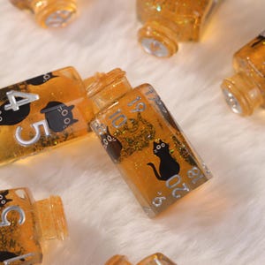 May include: Several small, clear, rectangular bottles filled with orange liquid and glitter. Each bottle has silver numbering and a black cat silhouette. The bottles are arranged on a white, fluffy surface.