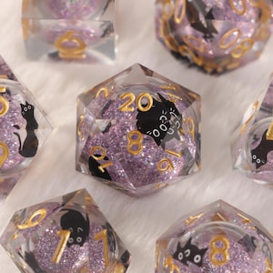 May include: Close-up of several polyhedral dice. The dice are transparent with a purple glitter interior and gold numbering. Each die features a black cat silhouette on some of the faces. The die showing the number 20 is in focus.