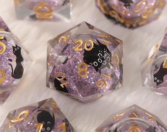 May include: Close-up of several polyhedral dice. The dice are transparent with a purple glitter interior and gold numbering. Each die features a black cat silhouette on some of the faces. The die showing the number 20 is in focus.