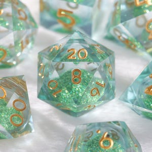Puede incluir: Primer plano de un conjunto de dados poliédricos. Los dados son transparentes con un tinte turquesa y numeración dorada. El interior de los dados tiene un relleno verde brillante. Los dados están diseñados para juegos de rol de mesa.
