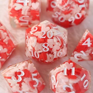 Peut inclure: Plusieurs dés polyédriques remplis de confettis en forme de cœur rouges et roses. Les dés présentent des chiffres blancs et sont de différentes formes, dont un d20, d10 et d4. Les dés sont posés sur un fond doux et flou.