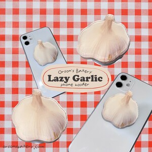 May include: A white phone case with a garlic-shaped phone holder. The phone holder is made of clear plastic and has a realistic garlic design. The text "Orson's Bakery Lazy Garlic phone holder" is printed on the case.