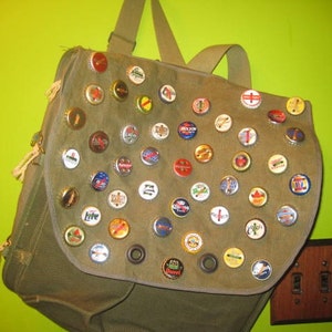 May include: A green canvas backpack with a variety of bottle caps attached to the front. The bottle caps are from different brands of beer and soda.