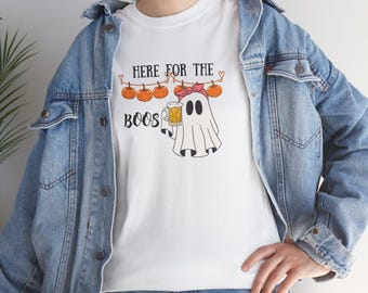 May include: A white t-shirt featuring a graphic of a ghost wearing a pink headband and holding a beer mug. Above the ghost, orange pumpkins hang from a string with the text "HERE FOR THE BOOS" printed in black. The shirt is worn under a blue denim jacket.