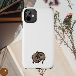 May include: A white smartphone case featuring a small, centered illustration of a brown, curled-up cat. The case has a black camera cutout at the top left corner, designed to fit a dual-lens camera system on a mobile device.