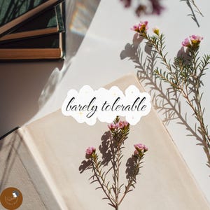 Könnte beinhalten: Ein weißer, konturgestanzter Aufkleber mit dem schwarzen kursiven Text "barely tolerable", der mit kleinen goldenen Sternchen verziert ist. Der Aufkleber liegt auf einem offenen Buch mit hellen Seiten, begleitet von Zweigen mit kleinen rosa Blüten.