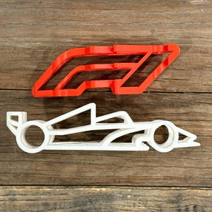 Puede incluir: Dos cortadores de galletas impresos en 3D: un logotipo de Fórmula 1 rojo y un coche de carreras blanco. El logotipo es una letra "F" estilizada con un contorno rojo. El coche de carreras es un contorno detallado de un vehículo de Fórmula 1.