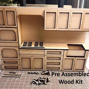 May include: A pre-assembled wood kit of a miniature kitchen. The kit includes a stove, sink, cabinets, and drawers. The kitchen is made of light-coloured wood and has a detailed design. The text "Pre Assembled Wood Kit" is visible.