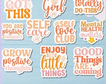 May include: A collection of inspirational stickers in shades of orange, pink, and white. The stickers feature phrases like "You Got This," "Self Care," and "Good Things Are Coming." The words are in a variety of fonts and styles, with floral accents.