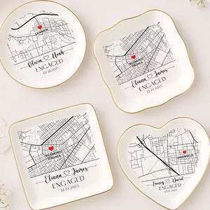 May include: Four white ceramic ring dishes with gold rims. Each dish features a black and white map design with a red heart marking a location. Text includes names and the word "ENGAGED" with a date. Shapes include round, square, and heart.