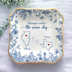 May include: A white ceramic dish with a gold rim, featuring blue floral patterns and the text "Always under the same sky." The dish includes state outlines with hearts and names, ideal for a personalized gift.