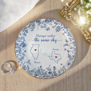 May include: A white decorative plate with a blue floral design and gold trim. The plate features the text "Always under the same sky" and illustrations of the states of Illinois and Texas, with the names "Olivia" and "Henry" below.