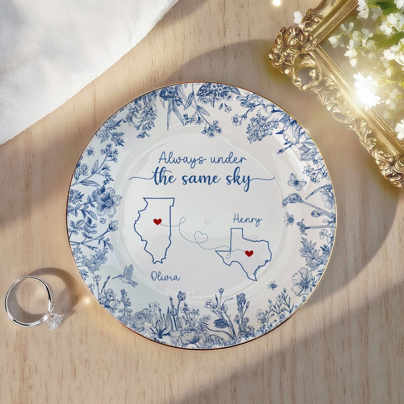 May include: A white decorative plate with a blue floral design and gold trim. The plate features the text "Always under the same sky" and illustrations of the states of Illinois and Texas, with the names "Olivia" and "Henry" below.