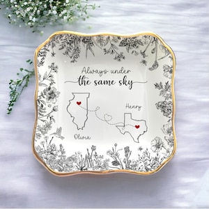 May include: A white ceramic trinket dish with a gold rim, featuring a floral design and the text "Always under the same sky." The dish includes illustrations of Illinois and Texas, connected by dotted lines and hearts.