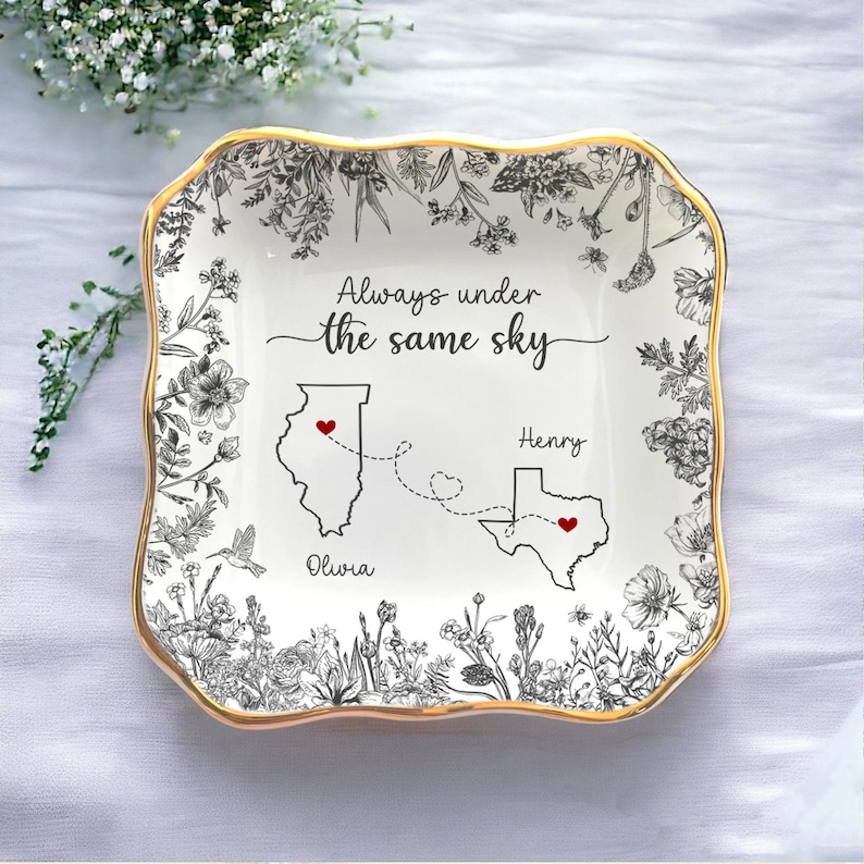 May include: A white ceramic trinket dish with a gold rim, featuring a floral design and the text "Always under the same sky." The dish includes illustrations of Illinois and Texas, connected by dotted lines and hearts.