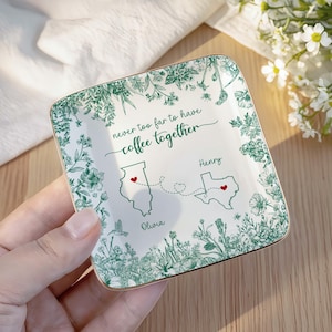 May include: A square, white ceramic trinket dish with a gold rim. The dish features a green floral border and the phrase "never too far to have coffee together." It includes state outlines with hearts and names.