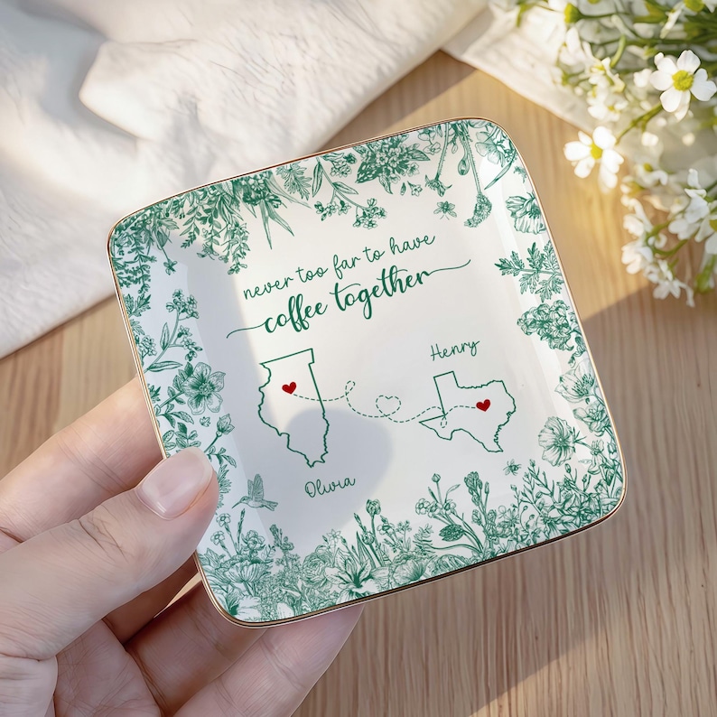 May include: A square, white ceramic trinket dish with a gold rim. The dish features a green floral border and the phrase "never too far to have coffee together." It includes state outlines with hearts and names.