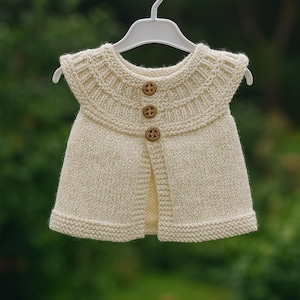 May include: Cream-coloured knitted baby vest with a round collar and three wooden buttons. The vest is hanging on a white hanger against a blurred green background. The knitted pattern is visible on the collar and hem.
