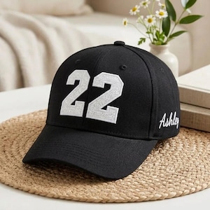 May include: Black baseball cap with the number "22" in white, embroidered on the front. The name "Ashley" is written in cursive on the side. The cap is sitting on a woven mat.