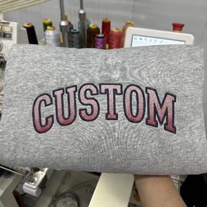 May include: A gray sweatshirt with the word "CUSTOM" embroidered in a gradient pink and red color. The sweatshirt is folded, and the embroidery is in a collegiate style font.