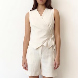 May include: A cream-colored sleeveless wrap top and matching shorts. The top has a V-neck and a tie closure at the waist. The shorts are high-waisted with a tailored fit. The outfit is made of a lightweight fabric.