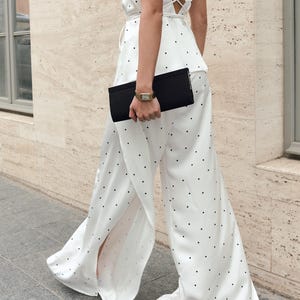 May include: A white satin jumpsuit featuring a black polka dot pattern, a halter neckline with a tie closure, an open back, and wide-leg trousers with side slits. The garment is paired with a rectangular black clutch bag and a gold-tone wristwatch.