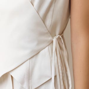 May include: Cream-colored wrap top with a tailored design. The top features a V-neckline and a wrap-around style that ties at the side with long, thin fabric ties. The fabric appears to be a medium-weight woven material.