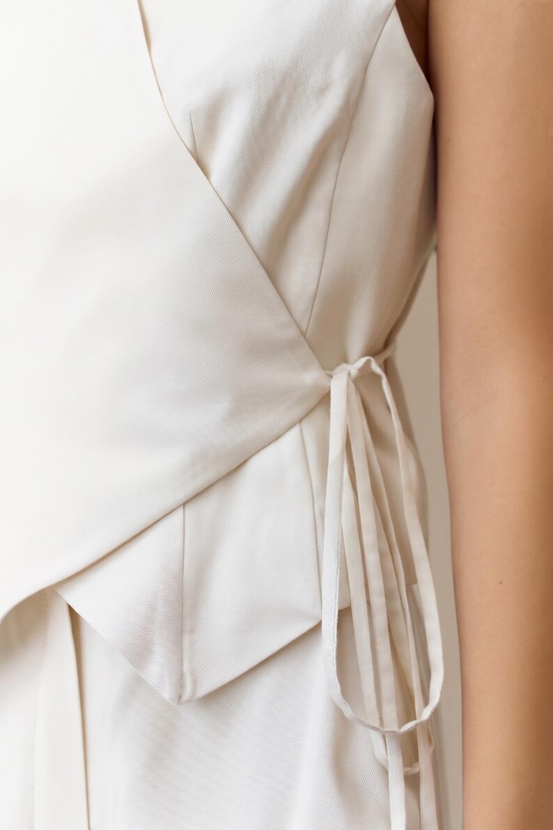 May include: Cream-colored wrap top with a tailored design. The top features a V-neckline and a wrap-around style that ties at the side with long, thin fabric ties. The fabric appears to be a medium-weight woven material.
